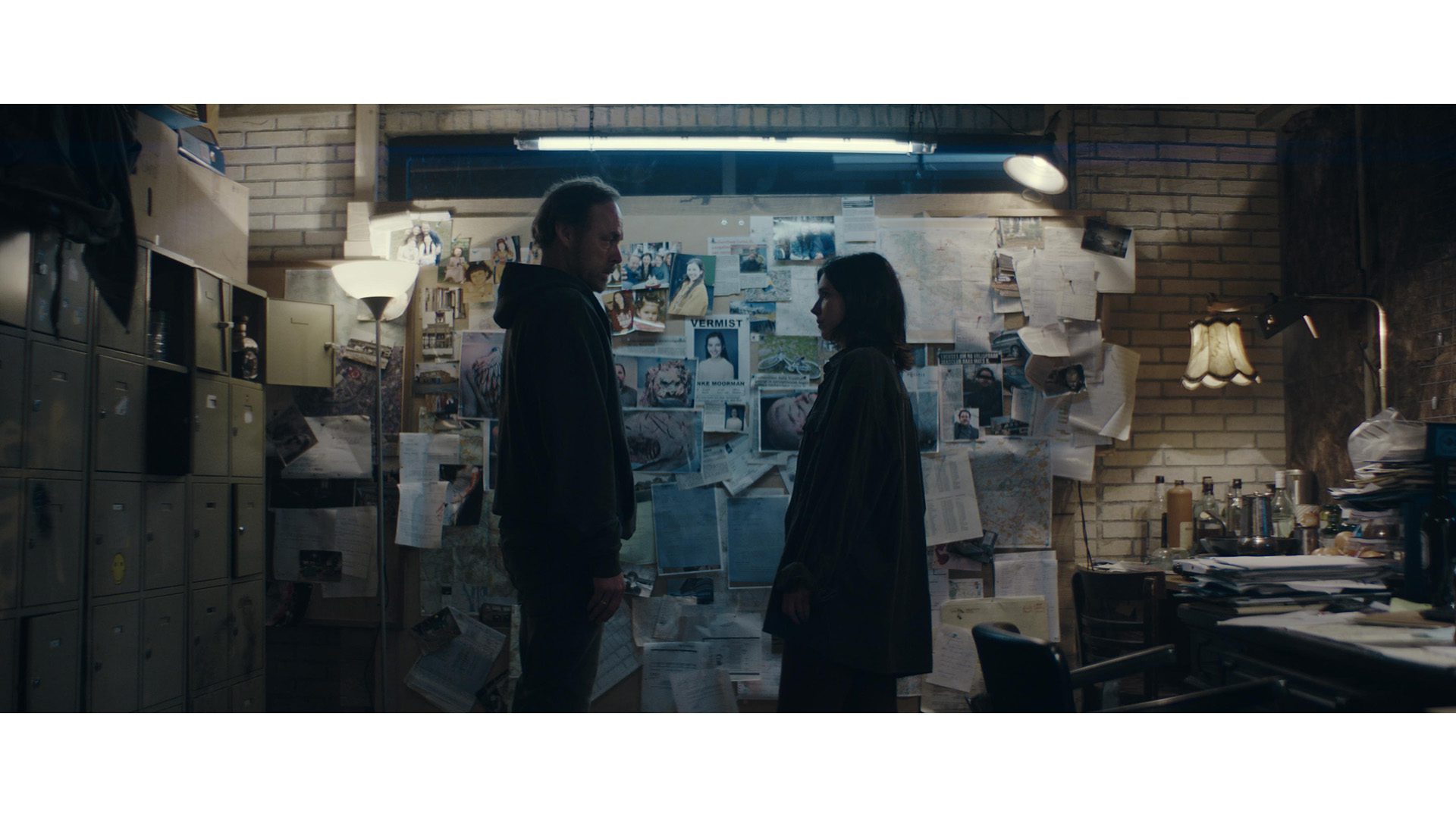Still from Sphinx. Minke and her dad are standing in front of a huge wall of newspaper clippings, photos, posters, etc. In the centre is the 'Missing' poster of Minke. Minke and her dad are seen in profile, with their faces to each other.