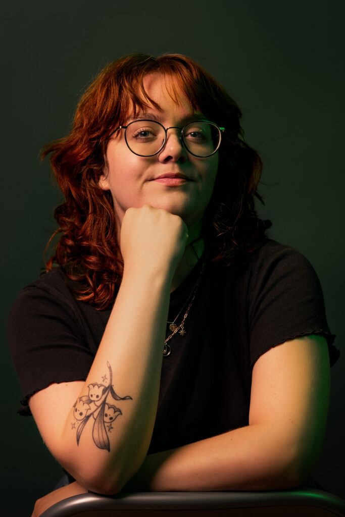 Portrait picture of me, Eva. I'm a white young woman with henna-dyed dark auburn wavy hair, cut in a wolf cut with lots of layers and blunt curtain bangs. I'm wearing dark green rimmed glasses, and I have my head leaning on my propped up right hand. On my right underarm is a tattoo visible of lily-of-the-valley flowers with little ghosts as the blooms. I'm sitting in front of a dark green background, and I'm wearing a black t-shirt, with two gold necklaces.