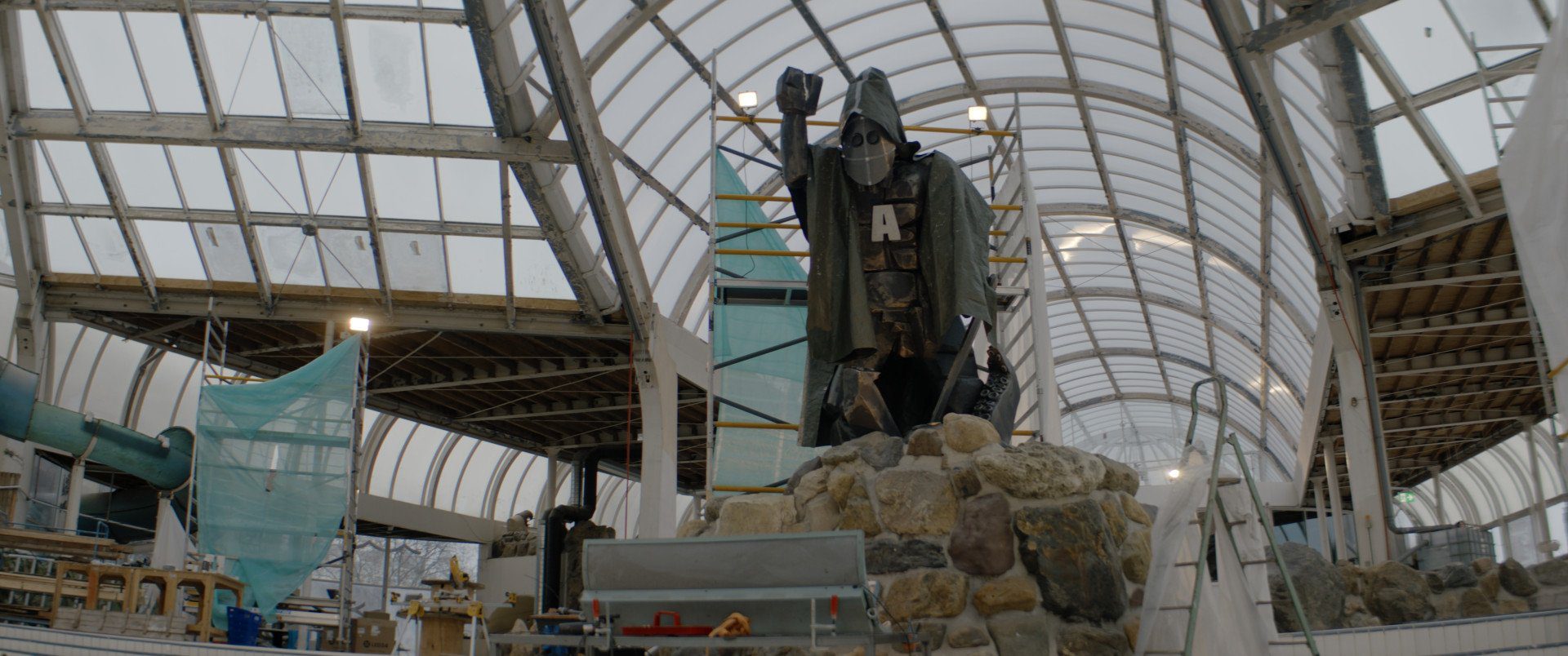 Still from Anoniem S2. A wide, low angle shot of a huge statue in a sort of abandoned swimming pool. The roof is a dome made up of glass panels and there are construction scaffolding strewn about. The statue is situated on fake rocks, has one hand lifted in the air, a mask on, a cape and a big white 'A' on it's chest.