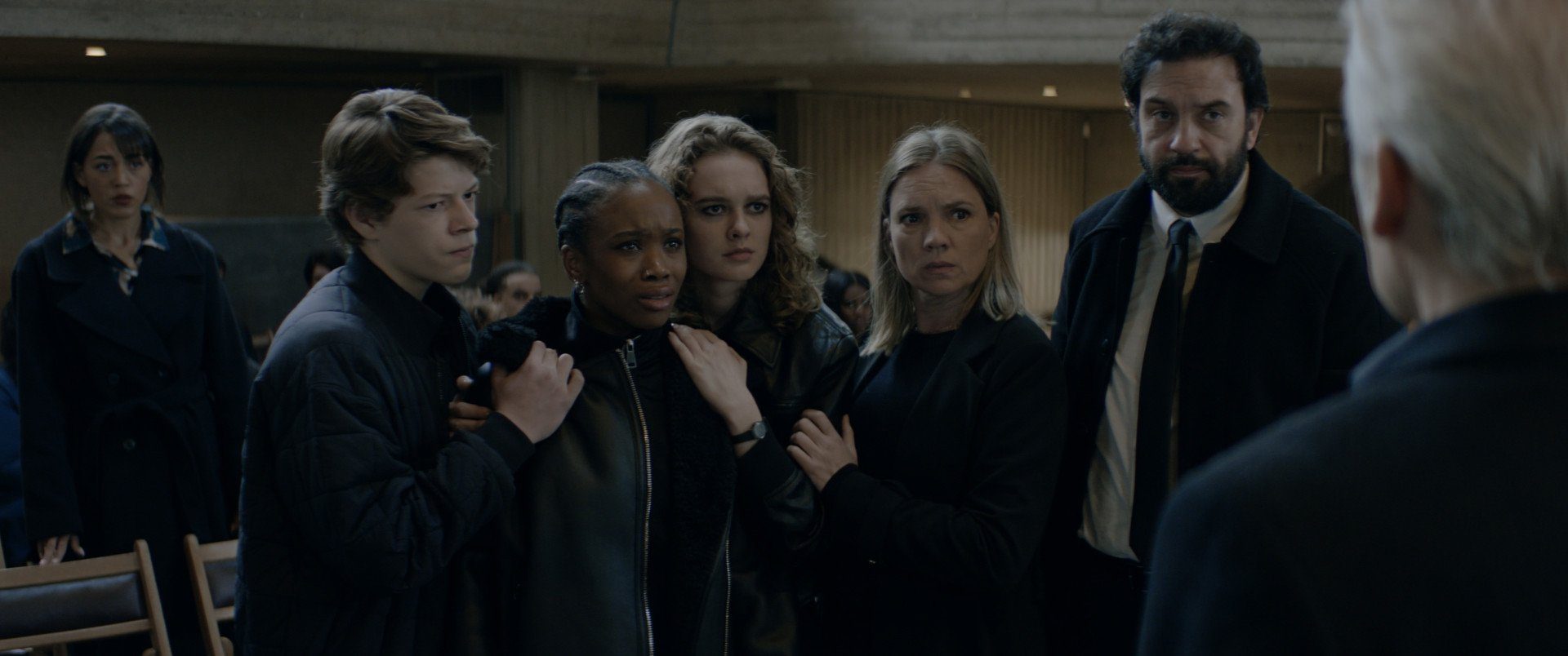 Still from Anoniem S2. We see a medium shot of the family, from left to right: Boet (Sil van der Zwan, a white 17-year-old boy looking angry), Mia (Xiorèse Romero, a black young adult woman with cornrows, with tears in her eyes), Ella (Mies van Rooijen, a white young adult woman with half long wavy blonde hair), Saar and Jurre (as previously described). In the background we see a couple of onlookers, and just seen over his shoulder is Cabo (Marcel Hensema).