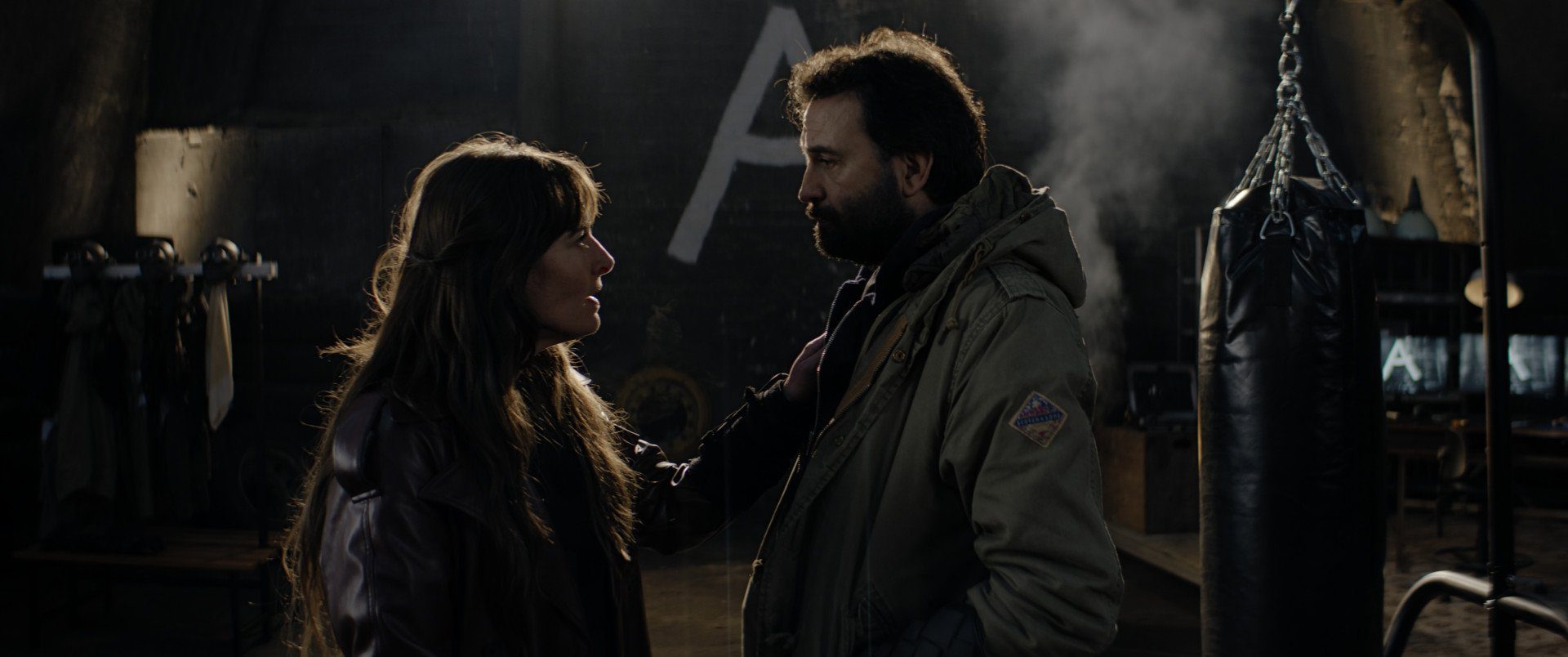 Still from Anoniem S2. An en-profil medium shot of Annet (Hannah van Lunteren, a white woman with long, brown hair with bangs) and Jurre (as previously described). They are standing in a non-descript dark room, with a big white 'A' on the wall behind them. They are looking at each other and Annet has her hand on Jurre's chest.