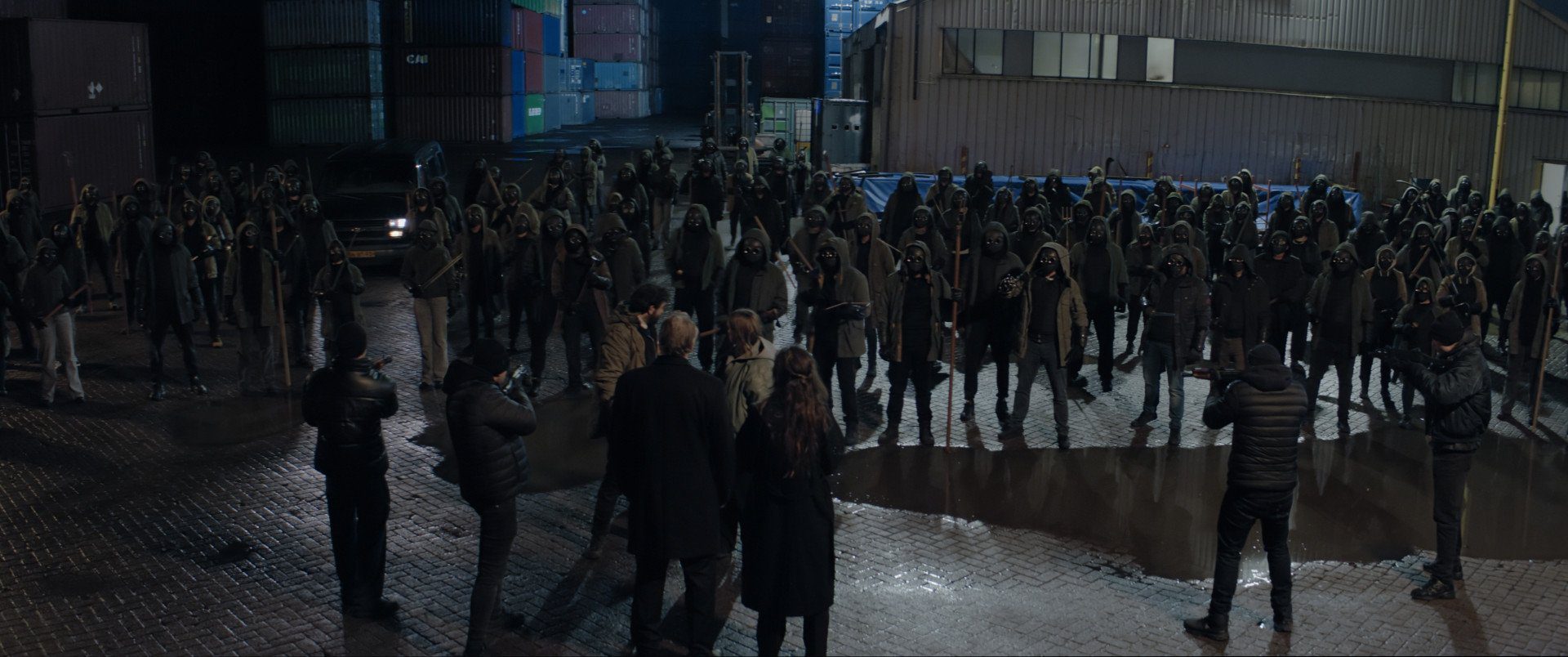 Still from Anoniem S2. A wide angle top shot of a group of people gathered in an industrial environment. We see a big group of anonymous 'Anoniem'-vigilantes. In front of them stand Jurre and Saar, both looking over their shoulder at the group. Facing them, with their backs to us, are 4 armed men, a middle-aged balding man and a woman with long brown hair.