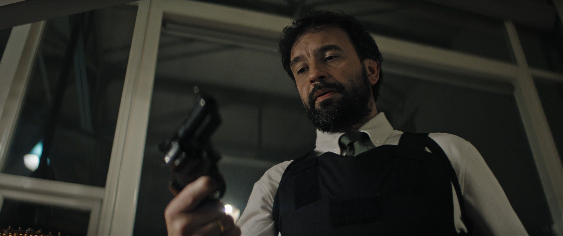 Still from Anoniem S2. A low angle medium shot of Jurre (Jeroen Spitzenberger), a middle-aged white man with dark hear and a dark, short beard. He is wearing a white button down with a tie under a kevlar vest and is looking down at a gun in his hand.