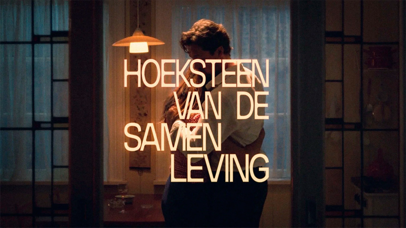 Title screen for Hoeksteen van de Samenleving. The image is a white, heterosexual couple in a 50s era kitchen at night. They are hugging closely. Overlaid, in a pale yellow font, is 'Hoeksteen van de Samenleving'.