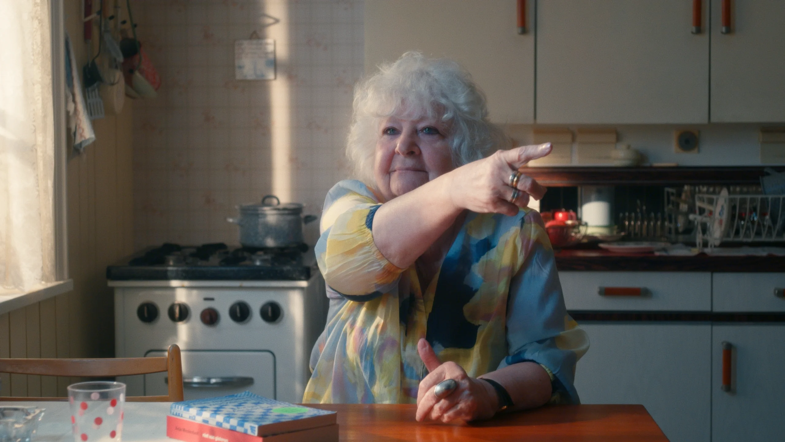 Still from Hoeksteen van de Samenleving. Writer and feminist Anja Meulenbelt, a white woman of about 80-years-old, is sitting in an old school kitchen at the kitchen table. She is wearing a blue and yellow floral blouse and is pointing to something/someone off screen.