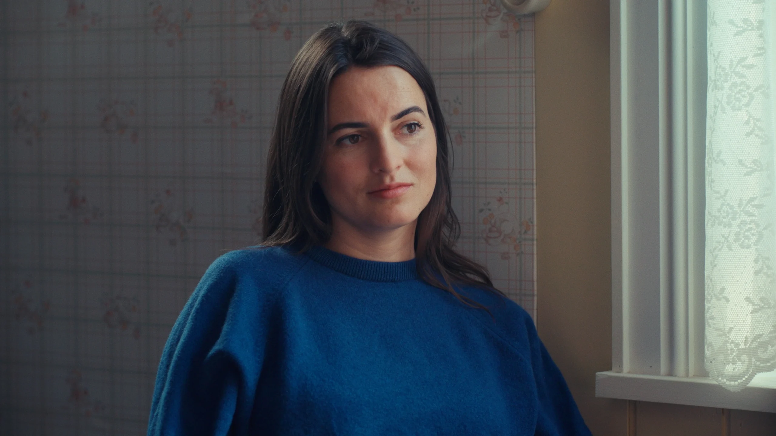 Still from Hoeksteen van de Samenleving. It's a medium-shot of director Juul Op den Kamp, who is listening intently to someone off screen. She is a white woman with long, dark hair and dark eyes. She is wearing a royal blue sweater and behind her is old school floral wallpaper.