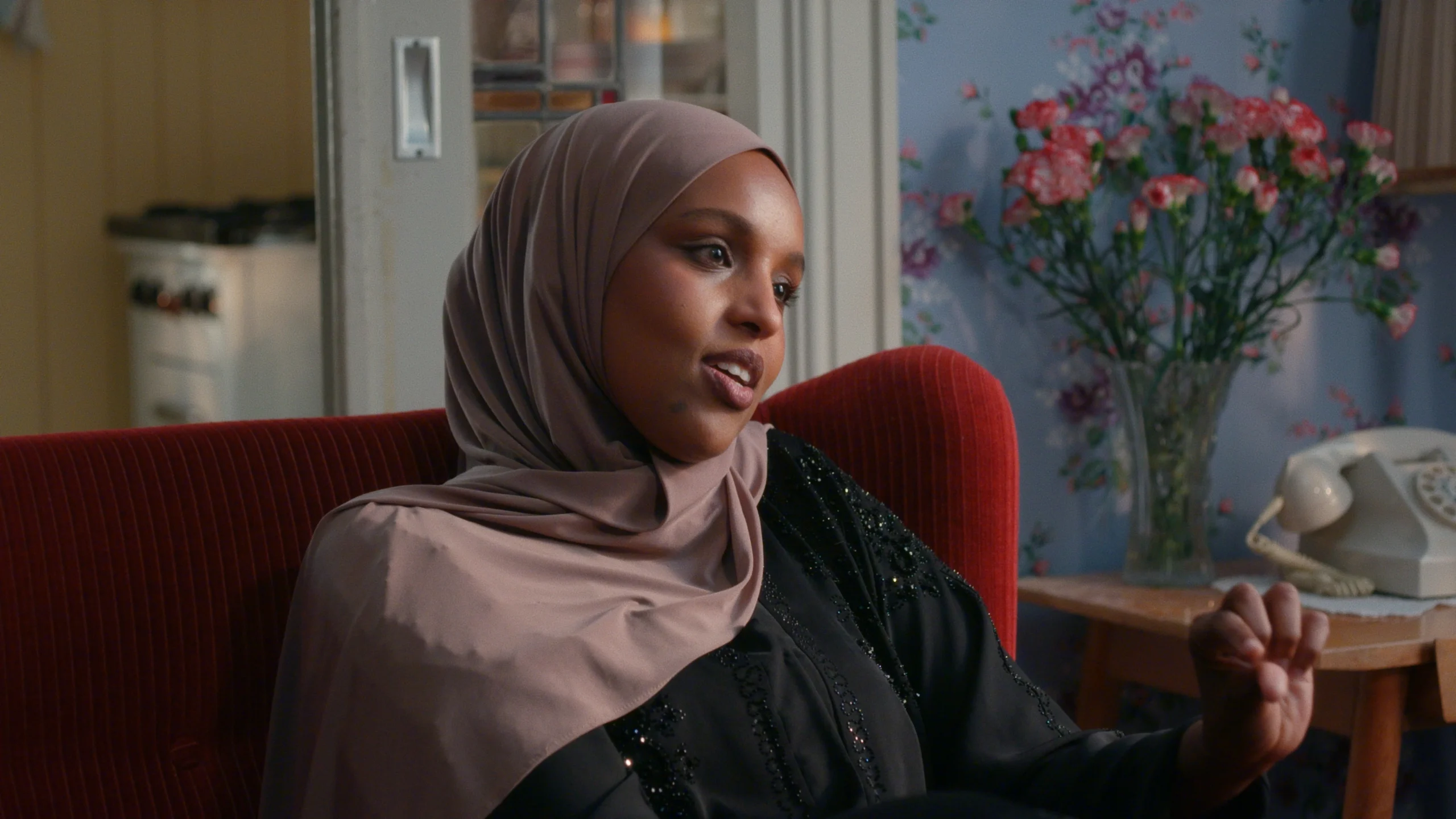 Still from Hoeksteen van de Samenleving. It's a medium close-up of Dr. Nawal Mustafa, a black woman wearing a lilac/beige hijab. She is sitting on a red couch in an old school living room with floral wallpaper.