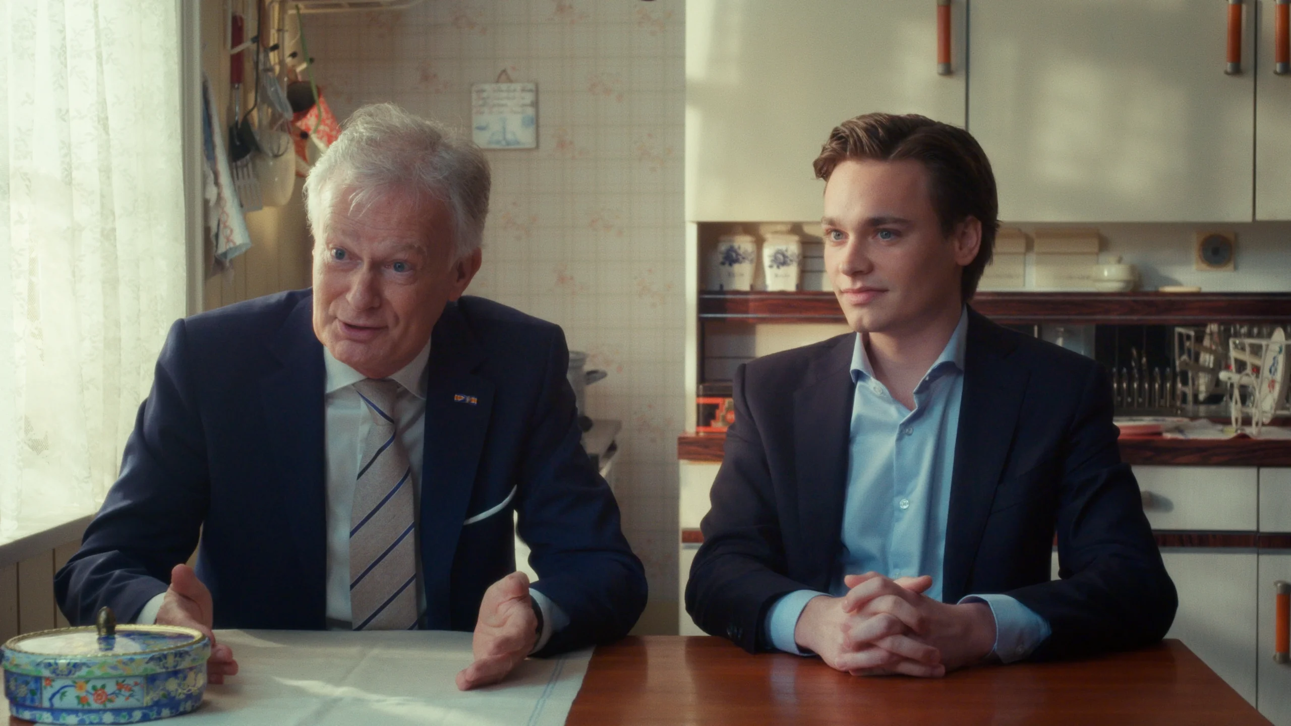Still from Hoeksteen van de Samenleving. It's a medium shot of Thomas Steenkamp and Thomas Steenkamp jr. Two white men, father and son, wearing neat suits, sitting at the kitchen table in a retro kitchen. The father is saying something, gesturing with his hands on the table.