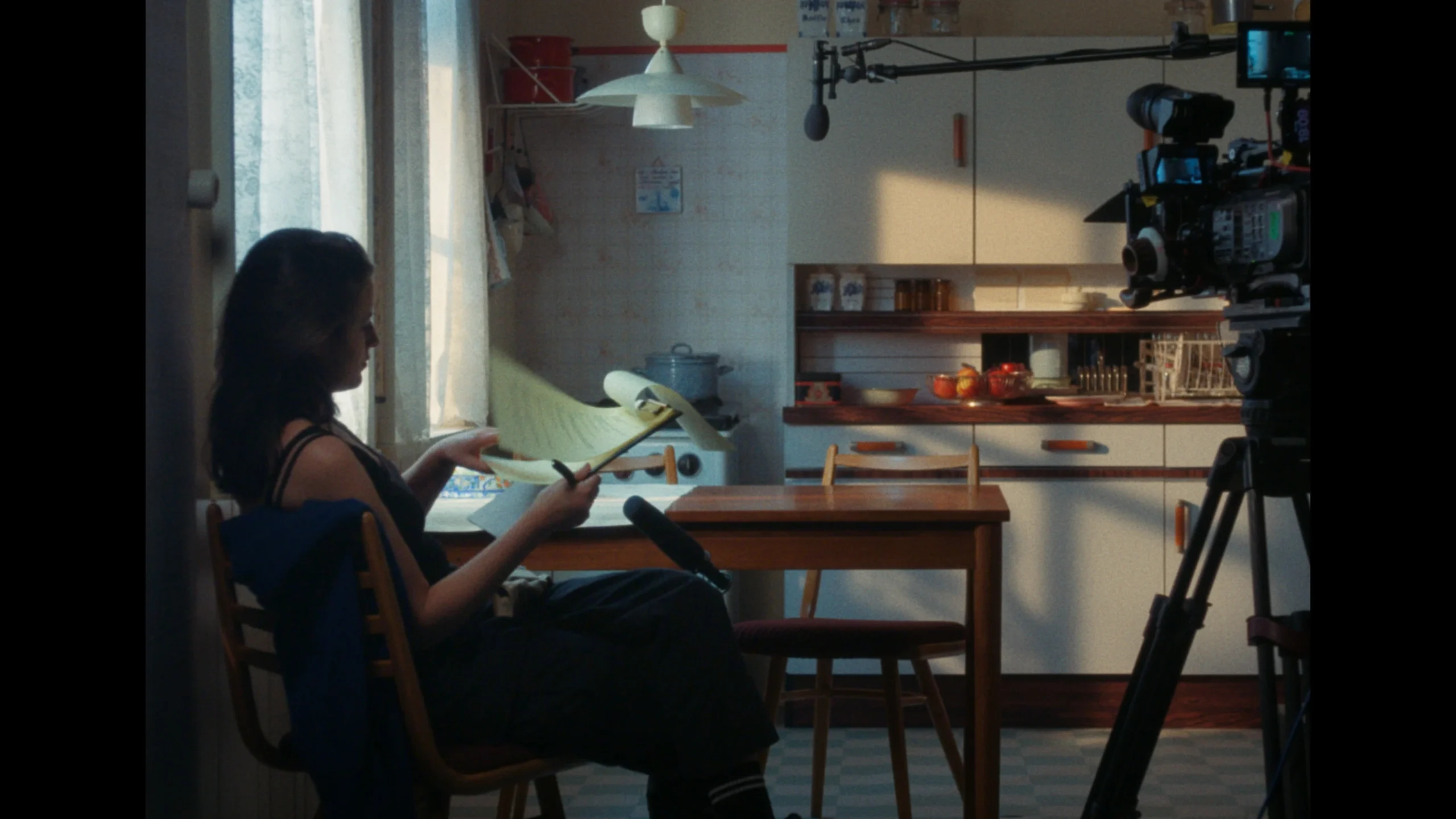 Still from Hoeksteen van de Samenleving. Wide shot of the retro kitchen, with a film camera on the right side and a boom mic in shot. Sitting at the side of the kitchen table closest to us is Juul Op den Kamp, reading her notes for the interview she is about to do.