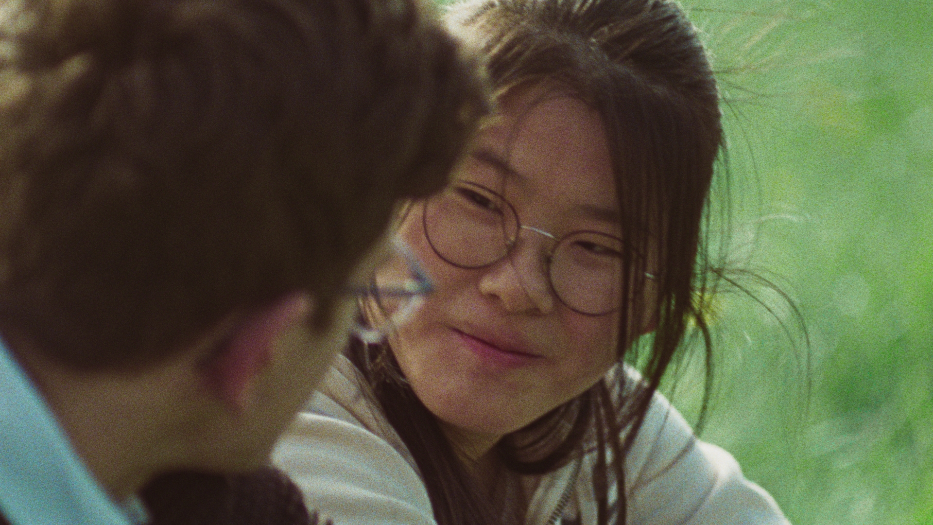 Still from 'Hier bij jou is alles wat ik wil'. It's a close-up over the shoulder shot on Caitlyn. She is a young Dutch-Asian girl of around 16-years-old with her long dark hair tied up in a ponytail. She is wearing round glasses and is looking lovingly at Abel, the 16-year-old white boy whose shoulder we are looking over.