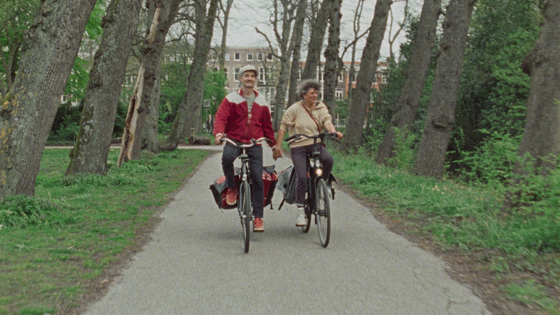 Still from 'Hier bij jou is alles wat ik wil'. A wide shot of Abel and Hester biking through Frankendaalpark.. They are holding hands.