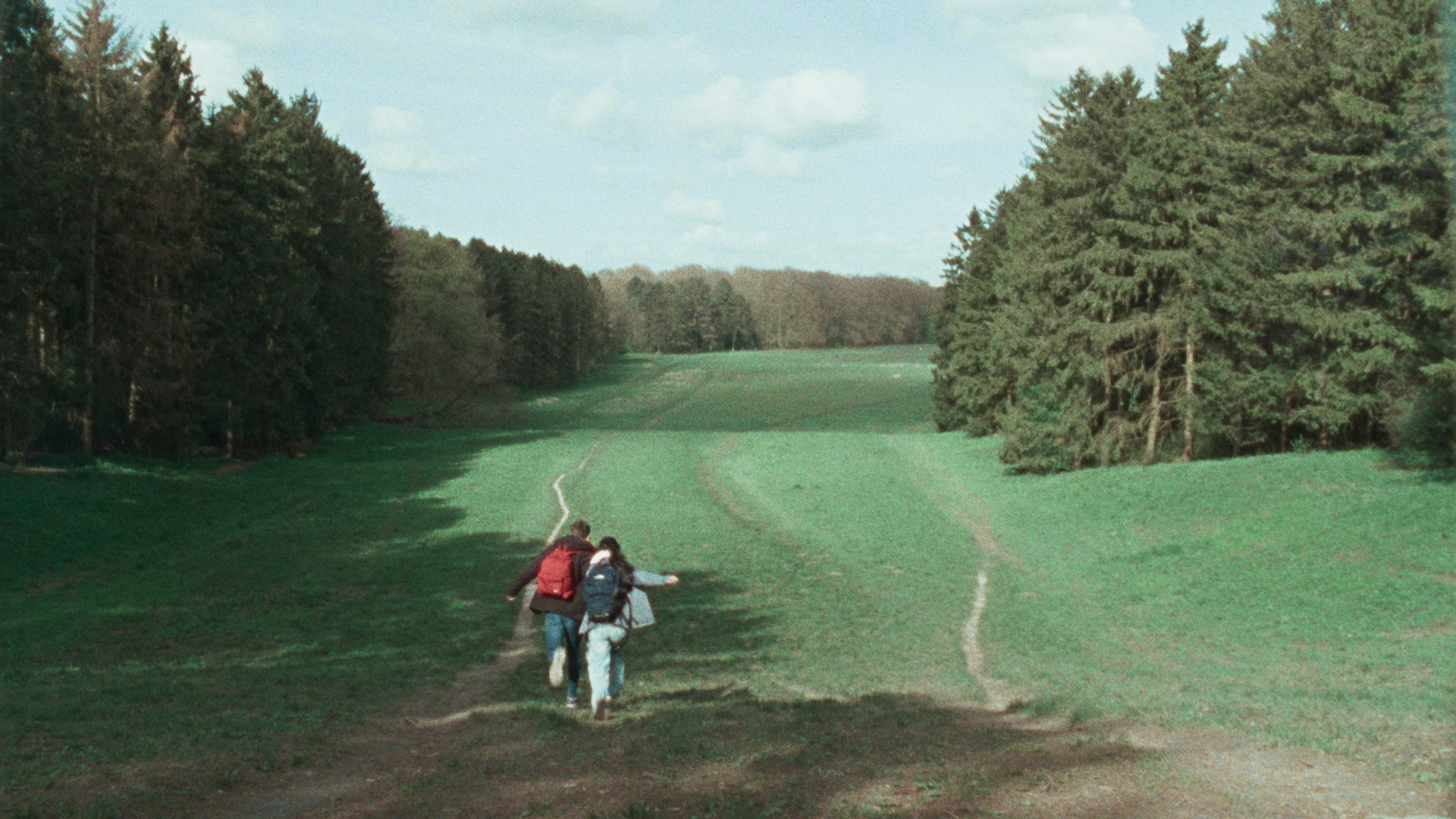 Still from 'Hier bij jou is alles wat ik wil'. A wide shot of (young) Abel and Caitlyn running away from us down a hill. They are both wearing backpacks, one red, one blue.