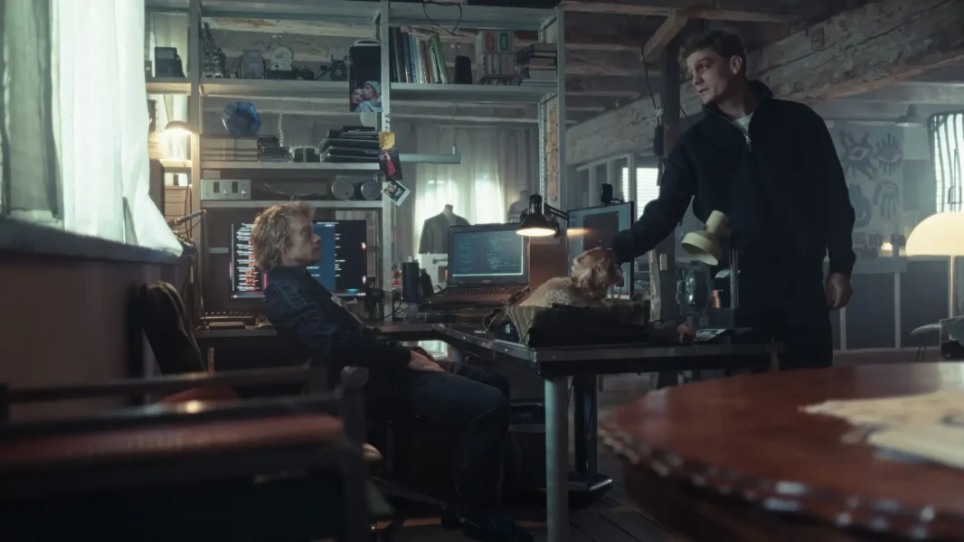 Still from Safe Harbor. Wide shot of Marco's apartment. Tobias (white, half long blonde hair) is sitting at a desk, leaning over. Marco (white male, short blonde hair) is standing on the other side of the desk, petting a dog that's laying on the desk.
