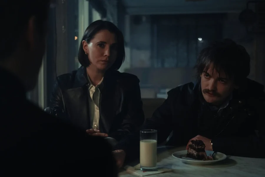 Still from Safe Harbor. Over-shoulder-shot on two people, Sloane and Farrell Walsch, are sitting at a table in a restaurant. Sloane is a white woman with a short, black bob. Farrell is a white man with messy brown half-long hair and a bushy moustache.