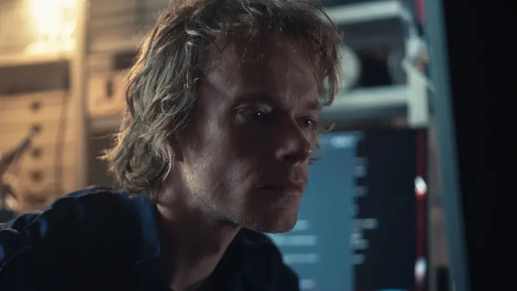Still from Safe Harbor. Close-up on Tobias, a white 30-something male with unkempt blonde half-long hair, is concentrating looking at a computer screen (off-screen).