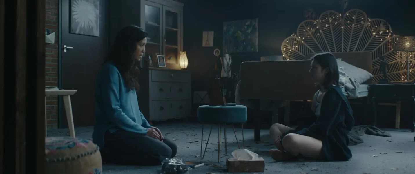 Still from Sphinx. This is a wide shot of Minke's bedroom. Minke is sitting cross legged on the floor on the right side of the frame, facing the left side. Across from her is an Asian woman with long black hair sitting on her knees. In between them is a stool with a metronome on it.